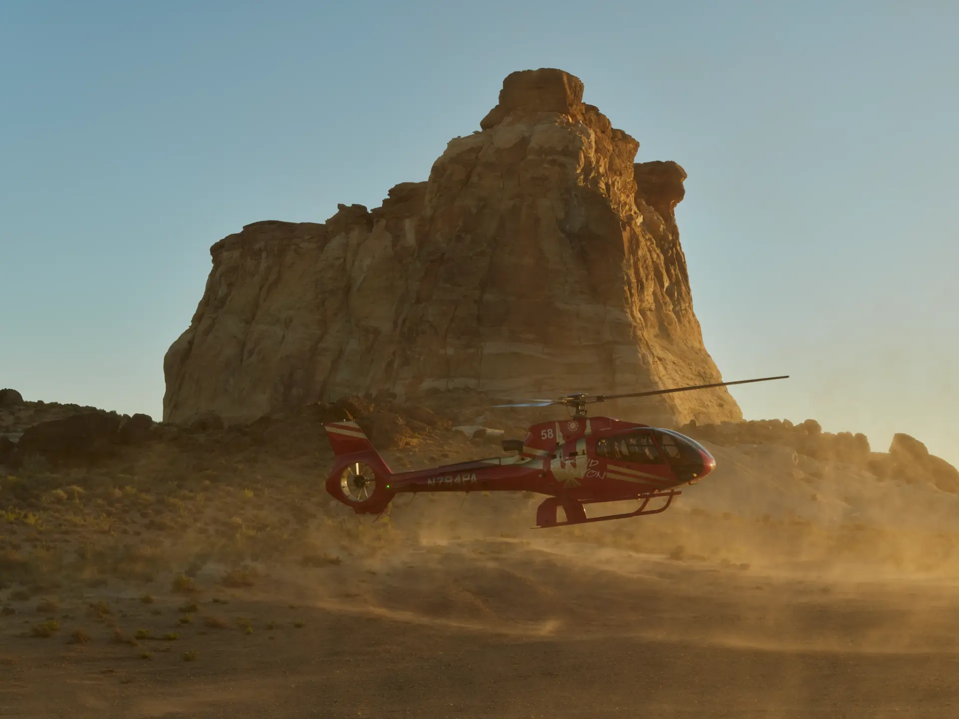 A red helicopter landing beneath a golden desert butte at dusk