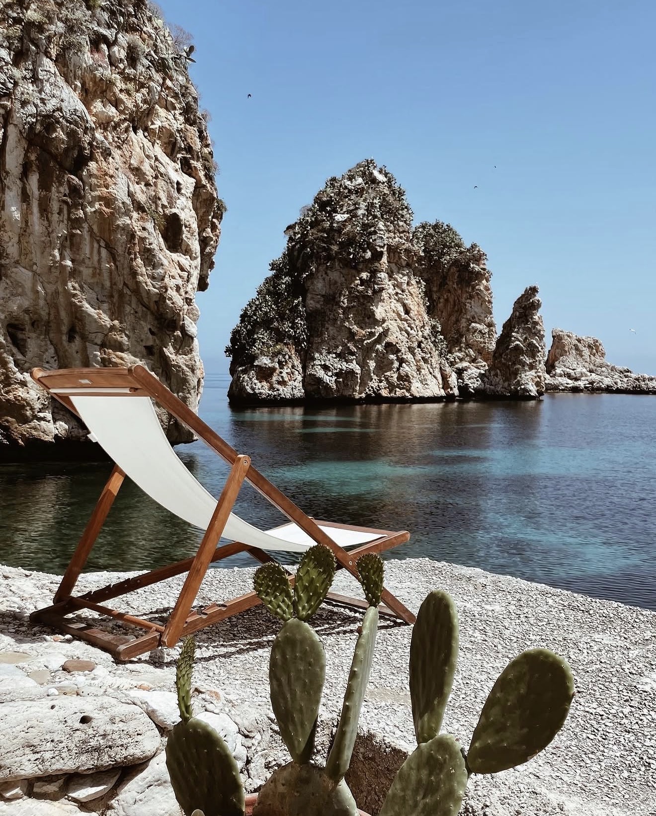 Teak deck chair on the stones at Scopello facing the faraglioni rocks and still sea