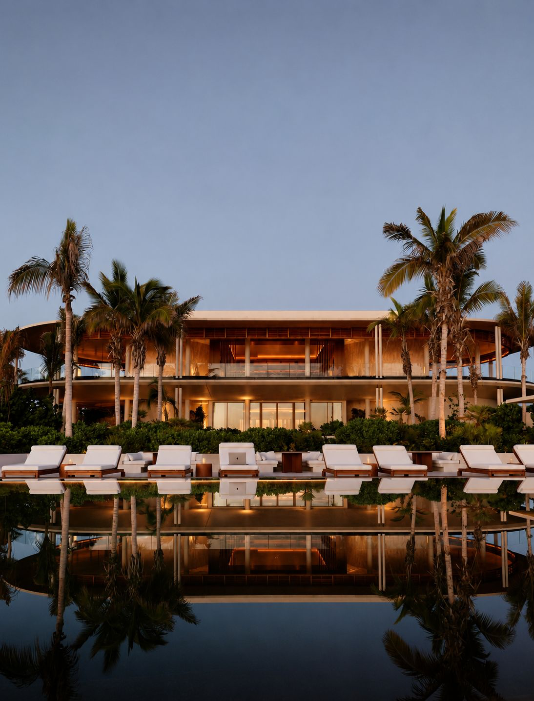 A contemporary villa at dusk, its warm light reflected in a still pool lined with palms and white daybeds
