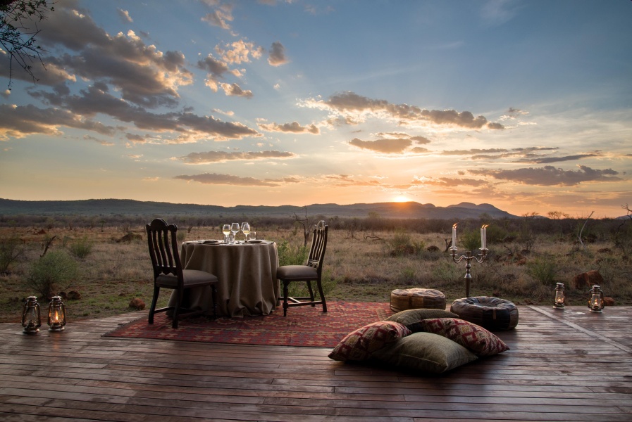 A candlelit safari lunch table set on a wooden deck at sunset, overlooking open plains